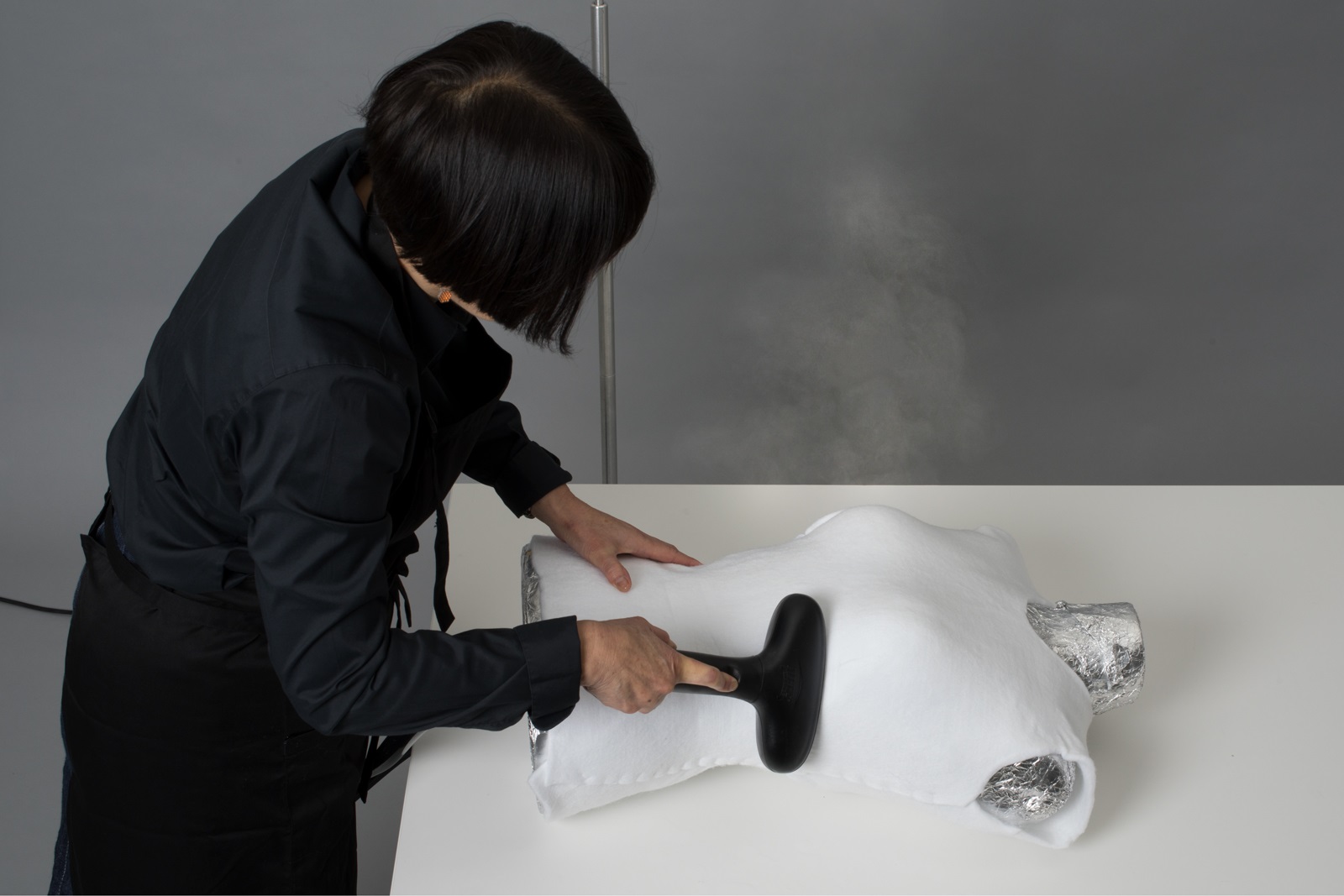 A woman works on a dress form lying on the table in front of her. The dress form is covered in aluminium foil over which Suzanne is steam-moulding a synthetic white felt in order to create a stylised body shape.